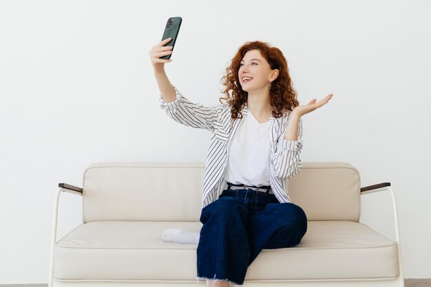 Glückliche lockige rothaarige Frau, die sich zu Hause auf einem Ledersofa ausruht und per Videoanruf mit der App auf dem Smartphone chattet, hat ein Gespräch mit der Familie, die Freunde grüßt und mit der Hand winkt