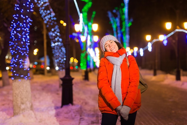 Glückliche lustige junge frau mit winterkleidung hintergrundabendlichterbeleuchtung der stadt. weihnachts- und winterferienkonzept.