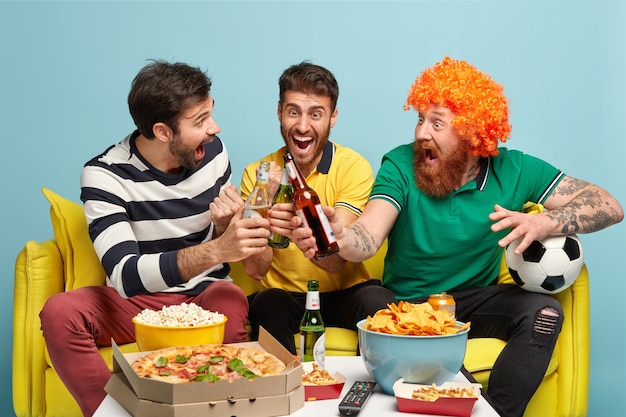 Glückliche Männer feiern den Sieg der Fußballmannschaft, die sie unterstützt haben, stoßen Bierflaschen an, schauen sich zu Hause ein Sportturnier an, essen einen Snack und schreien siegreich. Überglückliche Fans genießen den nationalen Wettbewerb im Fernsehen