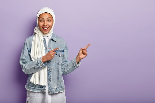 Glückliche Mischlingsfrau hat fröhlichen Ausdruck, zeigt mit beiden Vorderfingern weg, sagt dort folgen, zeigt Richtung über Kopierraum, trägt Jeanskleidung, weißen Hijab, isoliert auf lila Wand