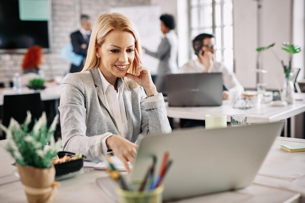 Glückliche mittlere erwachsene Geschäftsfrau, die Computer verwendet und im Internet surft, während sie im Büro arbeitet Es gibt Leute im Hintergrund