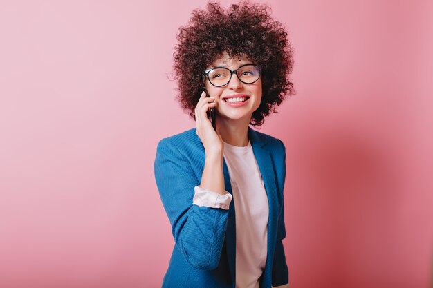 Glückliche moderne Frau trägt Brille und blaue Jacke, die am Telefon mit charmantem Lächeln auf Rosa sprechen