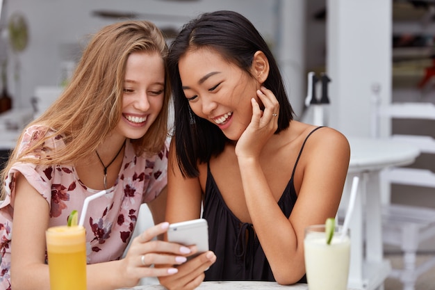 Glückliche multiethinische Frauen haben Spaß zusammen im Restaurant, lesen lustige Geschichten im Internet auf dem Handy, genießen frische kalte Getränke, haben eine gute Beziehung. Lächelnde freundliche Frauen mit Telefon