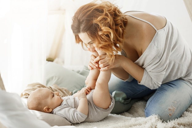 Glückliche Mutter, die mit neugeborenem Baby spielt, das kleine Beine küsst, die beste Mutterschaftsmomente im gemütlichen Schlafzimmer verbringen. Warme familiäre Atmosphäre. Familienkonzept.