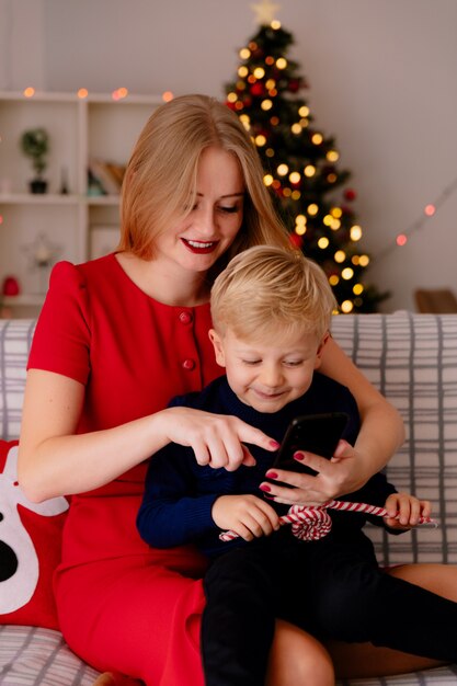 Glückliche Mutter im roten Kleid mit ihrem kleinen Kind, das auf einer Couch mit Smartphone in einem dekorierten Raum mit Weihnachtsbaum im Hintergrund sitzt