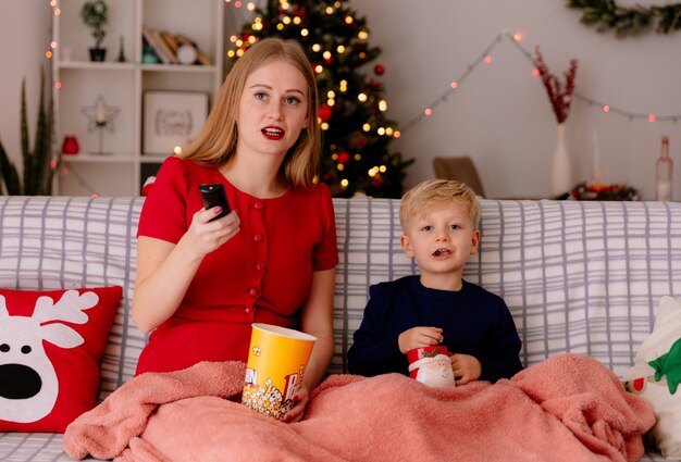 glückliche Mutter im roten Kleid mit ihrem kleinen Kind, das auf einer Couch unter Decke mit Eimer Popcorn sitzt, das zusammen in einem geschmückten Raum mit Weihnachtsbaum im Hintergrund fernsieht