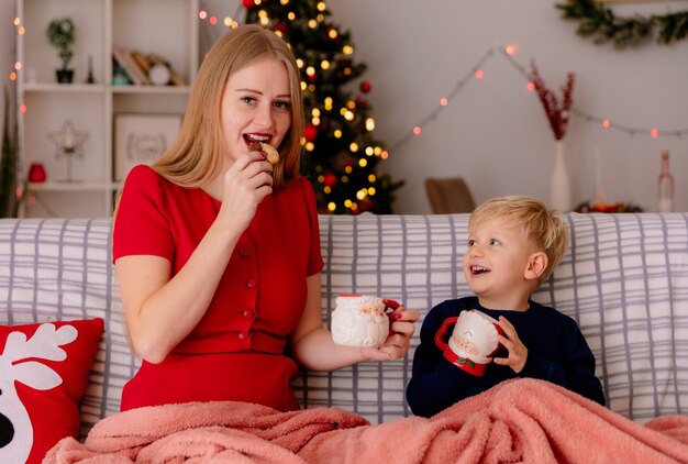 Glückliche Mutter im roten Kleid mit ihrem kleinen Kind, das auf einer Couch unter einer Decke sitzt und Tee aus Tassen trinkt und Kekse isst und in einem dekorierten Raum mit Weihnachtsbaum in der Wand lächelt