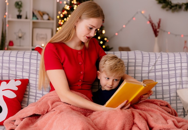 glückliche Mutter im roten Kleid mit ihrem kleinen Kind unter Decke Lesebuch in einem dekorierten Raum mit Weihnachtsbaum im Hintergrund