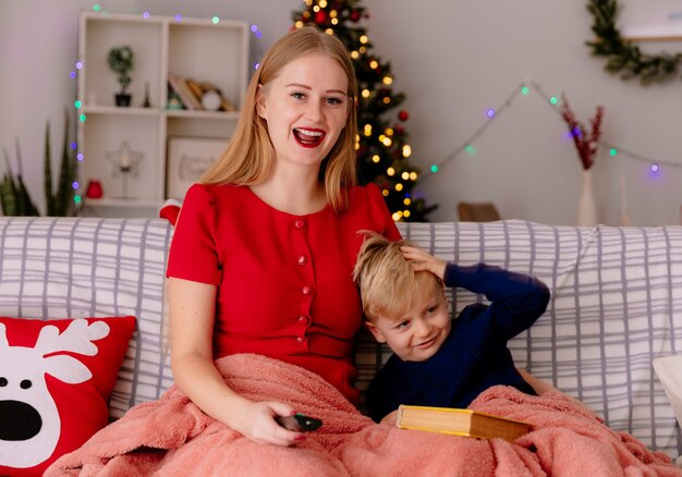 Glückliche Mutter im roten Kleid mit TV-Fernbedienung und ihr kleines Kind unter Decke mit einem Buch in einem dekorierten Raum mit Weihnachtsbaum in der Wand