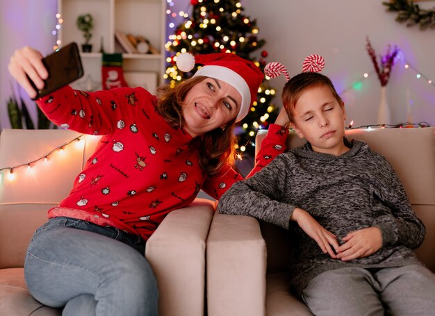 Glückliche Mutter in Weihnachtsmütze macht Selfie mit Smartphone mit ihrem kleinen Sohn, der auf einer Couch in einem dekorierten Zimmer mit Weihnachtsbaum in der Wand sitzt