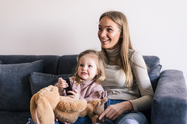 Glückliche Mutter mit der niedlichen kleinen Tochter, die Fernsehen mit Fernbedienung sieht