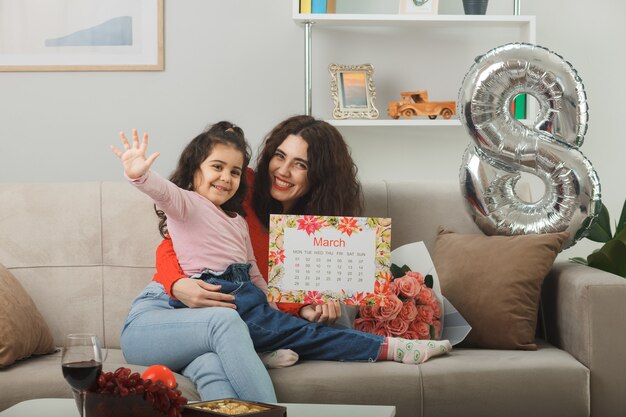 Glückliche Mutter mit ihrer kleinen Tochter, die auf einer Couch mit Blumenstrauß und Kalender des Monats März sitzt und fröhlich im hellen Wohnzimmer lächelt, um den internationalen Frauentag am 8. März zu feiern