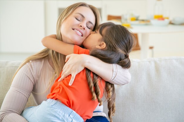 Glückliche Mutter sitzt mit ihrem kleinen Mädchen auf der Couch, hält Kind in den Armen und umarmt sie.