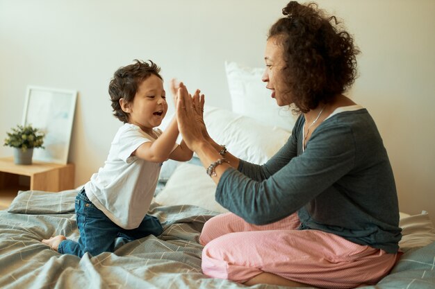 Glückliche Mutter und Baby spielen zu Hause. fröhliche lateinische Frau, die Hände mit ihrem kleinen Sohn fasst, der auf Bett sitzt