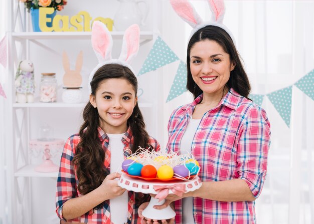 Glückliche Mutter und Tochter, die bunte Ostereier hält, nisten auf cakestand