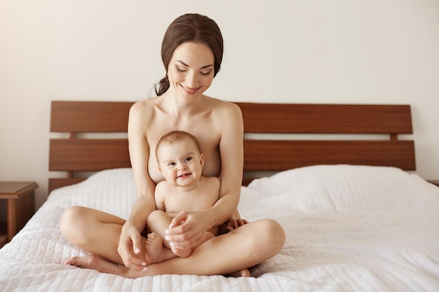 Glückliche nackte junge Mutter lächelt und umarmt ihr neugeborenes nettes Baby, das zusammen auf dem Bett sitzt
