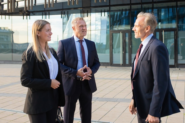 Glückliche positive Geschäftspartner treffen sich im Bürogebäude, stehen und reden im Freien und besprechen den Deal. Mittlere Einstellung, Seitenansicht. Kooperationskonzept