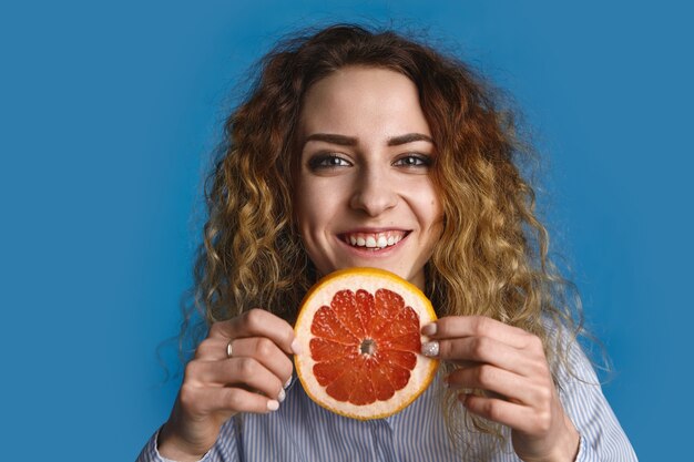 Glückliche positive junge 25-jährige Frau mit gewellter Frisur, die Scheibe der frischen Grapefruit hält und Hände ausstreckt, als ob Sie saftige vitaminreiche Frucht anbieten. Konzept für gesunden Lebensstil und Obstbau