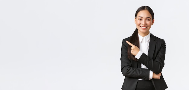 Glückliche professionelle asiatische Managerin, Geschäftsfrau im Anzug, die Ankündigung zeigt, lächelt und mit dem Finger auf das Produkt- oder Projektbanner zeigt, stehend auf weißem Hintergrund