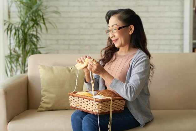 Glückliche reife strickende Kleidung der Asiatin, die zu Hause auf Sofa sitzt