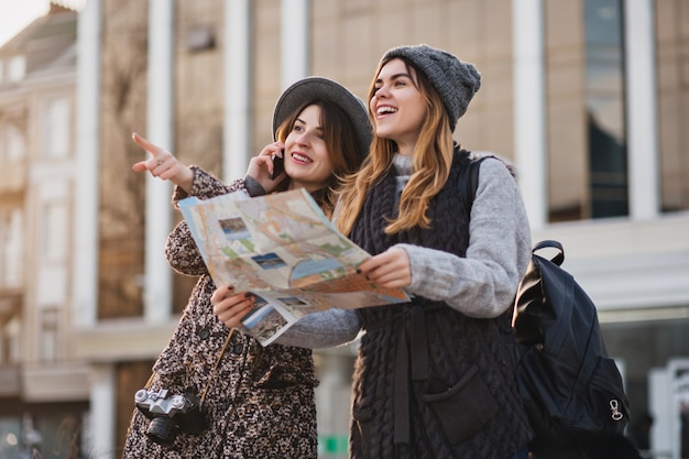 Glückliche Reise zusammen von zwei modischen Frauen im sonnigen Stadtzentrum. Junge freudige Frauen, die Positivität ausdrücken, Karte verwenden, Urlaub mit Taschen, fröhliche Gefühle, guten Tag.