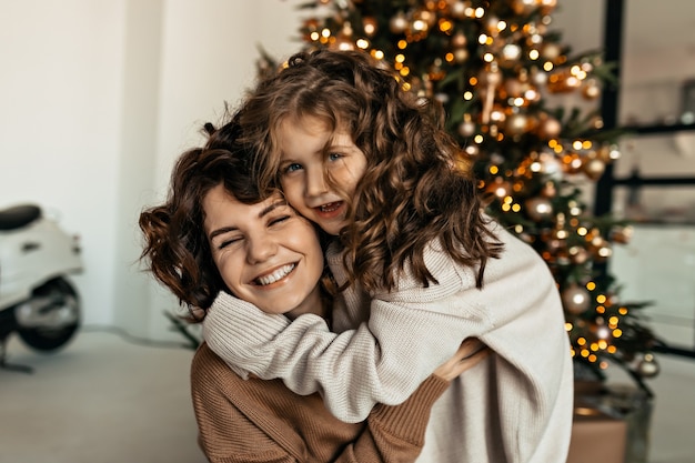 Glückliche reizende Frau mit ihrer kleinen süßen Tochter mit welligem Haar, das Spaß vor Weihnachtsbaum umarmt und Spaß macht