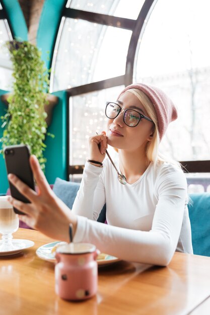 Glückliche reizende junge Frau im Hut und in den Gläsern, die essen und Selfie mit Smartphone im Café machen