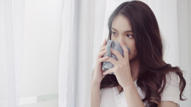Glückliche schöne Asiatin, die einen Tasse Kaffee oder einen Tee nahe dem Fenster im Schlafzimmer lächelt und trinkt.