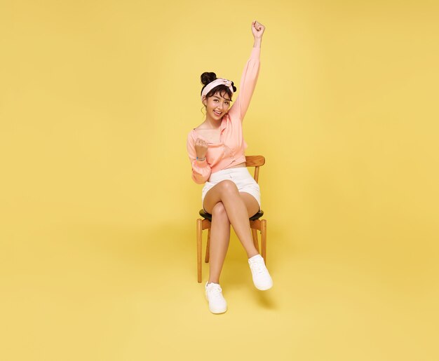 Glückliche schöne asiatische jugendlich Frau, die auf Stuhl sitzt und Hände hoch feiert Geste auf gelber Wand. Aufgeregtes Gewinner-Erfolgskonzept.