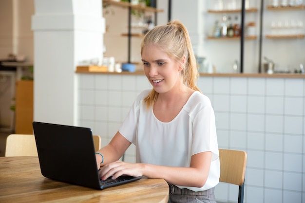 Glückliche schöne blonde Frau, die am Tisch im Co-Arbeitsraum sitzt, Laptop verwendet, Anzeige betrachtet und lächelt