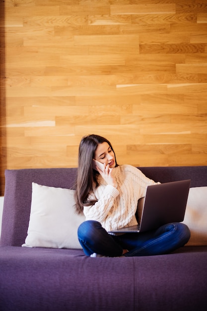 Glückliche schöne Frau, die an einem Laptop arbeitet, der auf dem Bett im Haus sitzt