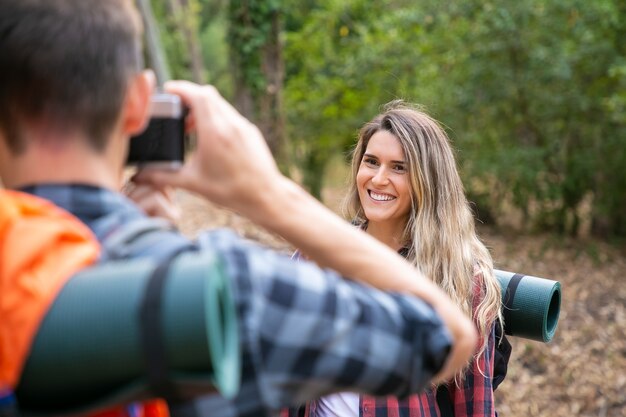 Glückliche schöne Frau, die für Foto aufwirft und auf Straße im Wald steht. Unscharfer Kerl, der Foto des kaukasischen lächelnden weiblichen Reisenden macht. Backpacking Tourismus, Abenteuer und Sommerurlaub Konzept