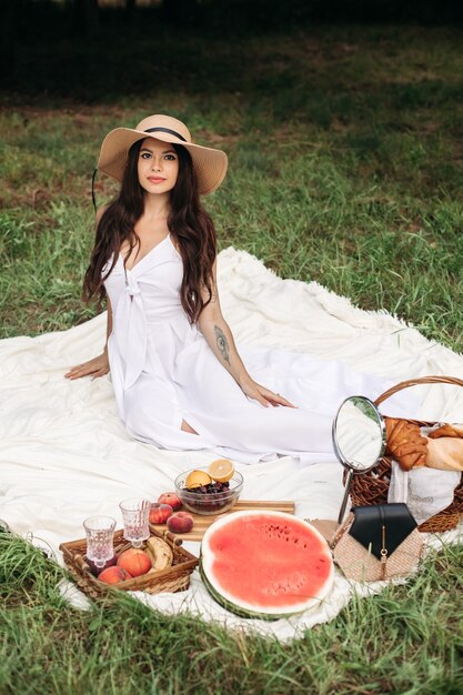 Glückliche schöne Frau mit Hut mit Krempe und weißem Kleid, während sie im Sommerpark steht und einen Korb mit Brot hält
