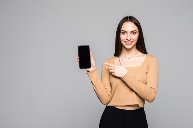 Glückliche schöne junge Frau, die Handy mit leerem Bildschirm über grauer Wand hält