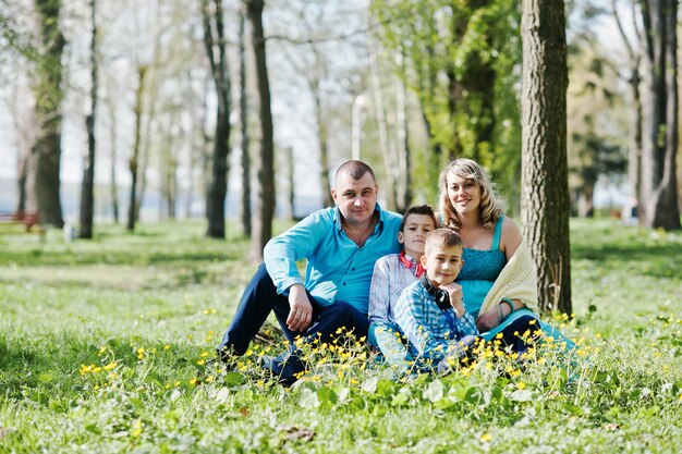 Glückliche schwangere Familie mit zwei Söhnen in türkisfarbenen Kleidern, die auf Gras mit Blumen im Park sitzen
