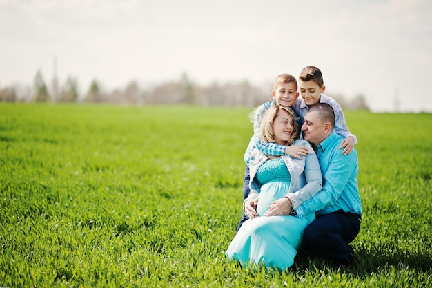 Glückliche schwangere Familie mit zwei Söhnen in türkisfarbener Kleidung, die gemeinsam Zeit auf grünem Gras auf dem Feld verbringen