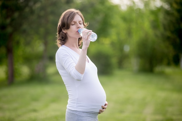 Glückliche schwangere Frau trinkt natürliches Wasser im Park