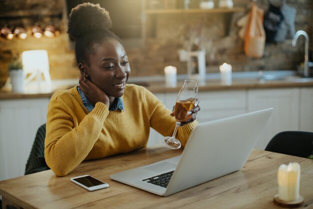 Glückliche schwarze Frau, die während des Videoanrufs zu Hause mit Champagner anstößt