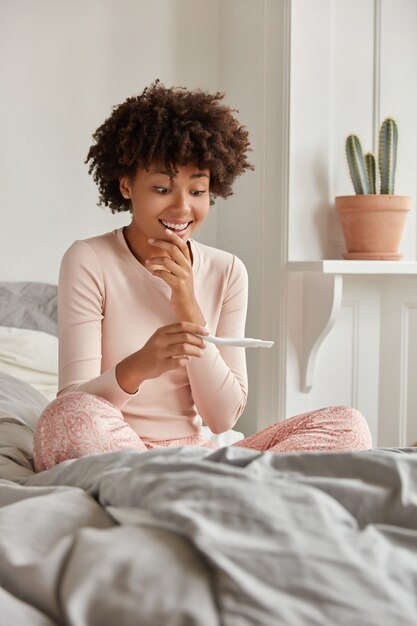 Glückliche schwarze Frau mit Afro-Frisur, sieht positiv auf Schwangerschaftstest aus