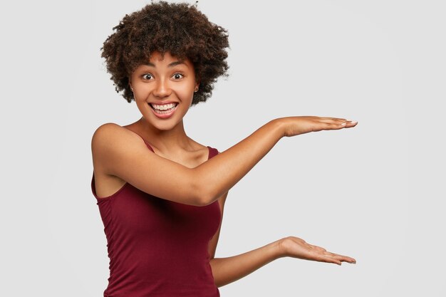 Glückliche schwarze junge Frau mit erfreutem Gesichtsausdruck zeigt Höhe von etwas