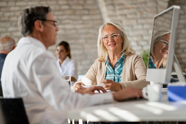 Glückliche Seniorin, die mit dem Gesundheitsdienstleister kommuniziert, während er medizinische Daten auf einem Computer in der Klinik analysiert