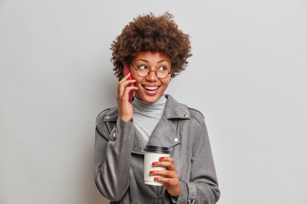 Glückliche sorglose junge Frau hat Afro-Frisur, genießt lebhafte Telefongespräche, schaut weg, hält eine Tasse Kaffee in der Hand, trägt eine runde Brille und eine graue Jacke