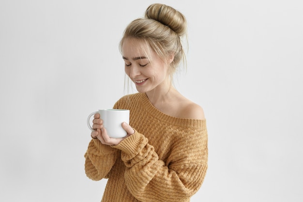 Glückliche sorglose junge Frau mit Haarknoten, die zu Hause nach der Arbeit breit lächelnd breit lächelt und guten Kaffee von der großen Tasse genießt. Attraktive Frau gekleidet in gemütlichen warmen Pullover trinken Kräutertee