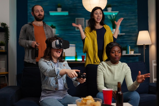 Glückliche Spielerfrau mit Virtual-Reality-Headset, die Online-Wettbewerbe gewinnt, die Videospiele mit Gaming-Joystick spielen und den Sieg mit Freunden zu Hause genießen. Konzept der multiethnischen Freunde, die rumhängen