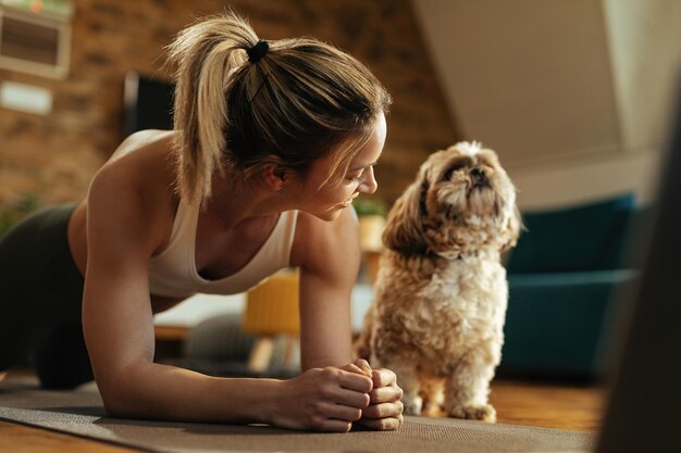 Glückliche sportliche Frau, die Plankenübungen macht, während ihr Hund neben ihr sitzt.