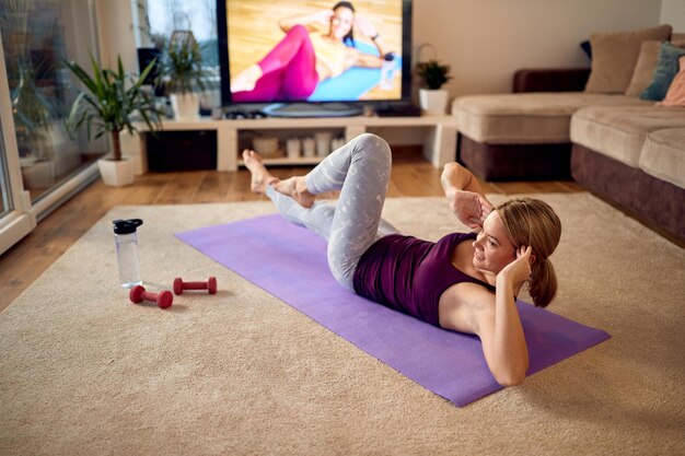 Glückliche sportliche Frau, die Situps praktiziert, während sie zu Hause vor einem Fernseher trainiert