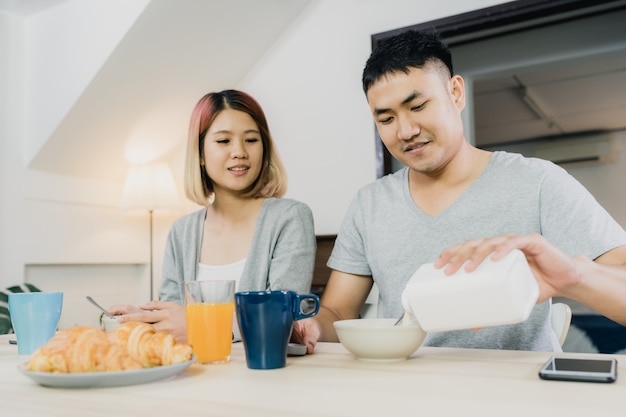 Glückliche süße asiatische Paare, die frühstücken, Getreide in der Milch, Brot und Orangensaft trinken