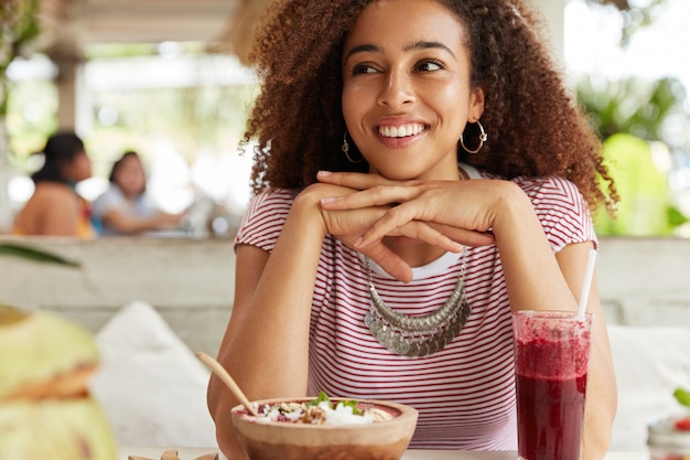 Glückliche süße Frau mit dunkler Haut, Afro-Frisur, schaut mit fröhlichem Ausdruck weg, hält Hände unter dem Kinn, isst köstliches exotisches Dessert und trinkt Cocktail, träumt von etwas Angenehmem