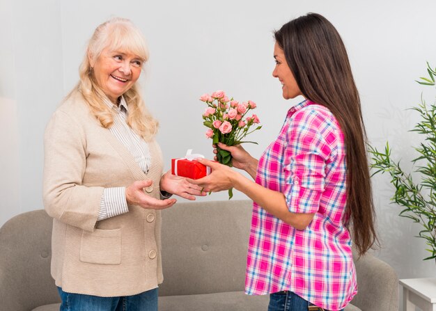 Glückliche Tochter, die ihrer Mutter eingewickelten Geschenkbox- und Blumenblumenstrauß gibt