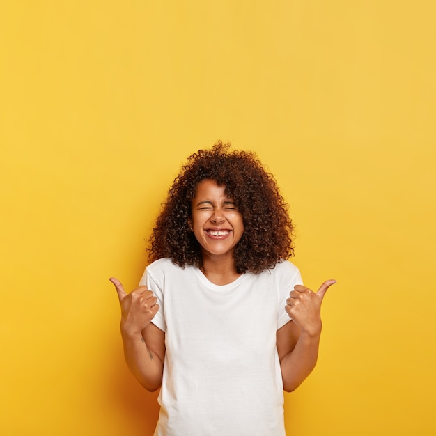 Glückliche überglückliche ethnische Dame mit buschigem, knackigem Haar gibt positive Antwort mit Daumen hoch, mag tolle Idee, schließt die Augen vor Lachen, gekleidet in Modell-T-Shirt, isoliert auf gelber Wand.